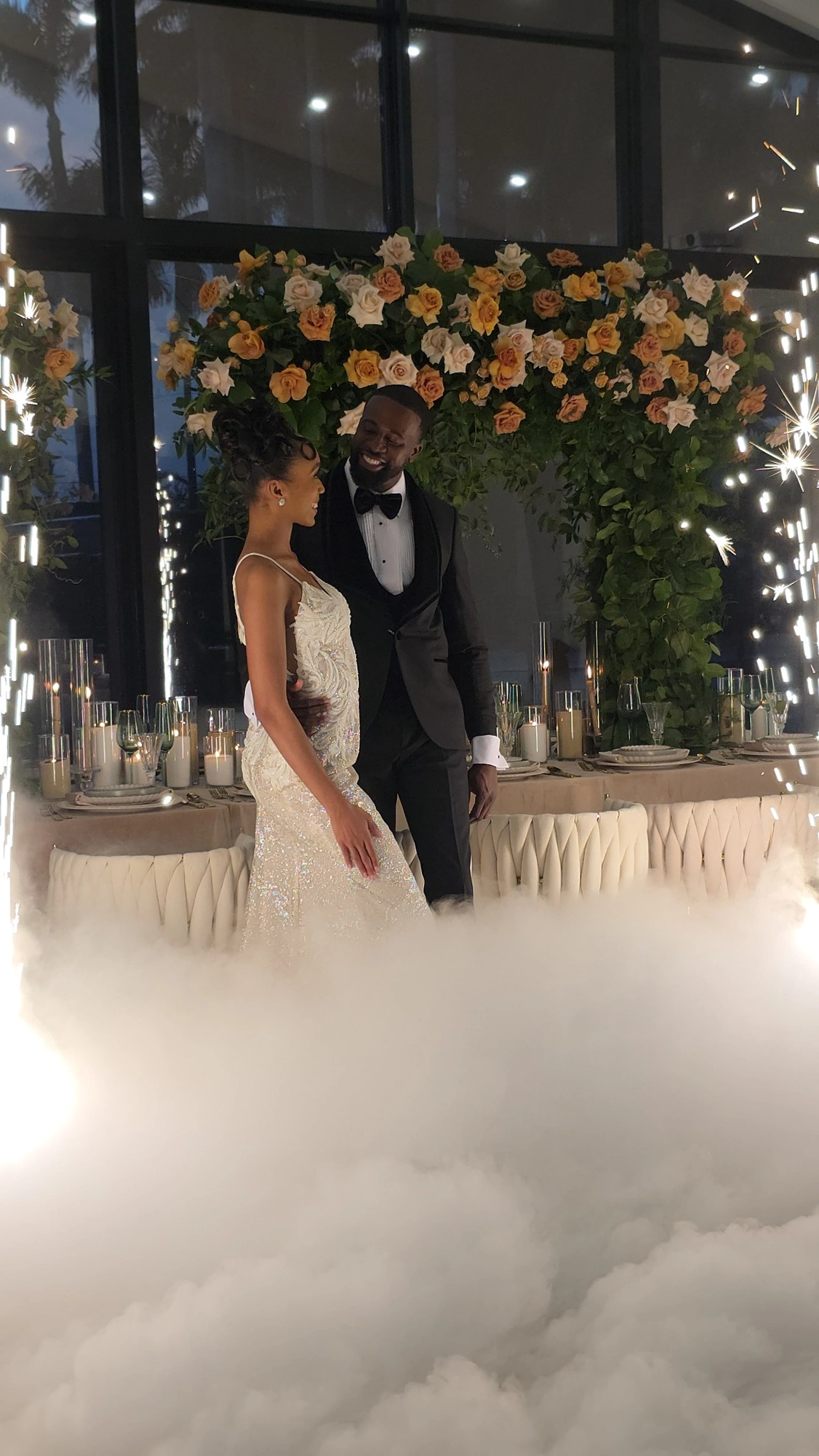 A couple sharing a joyful moment surrounded by florals, low fog, and spark effects