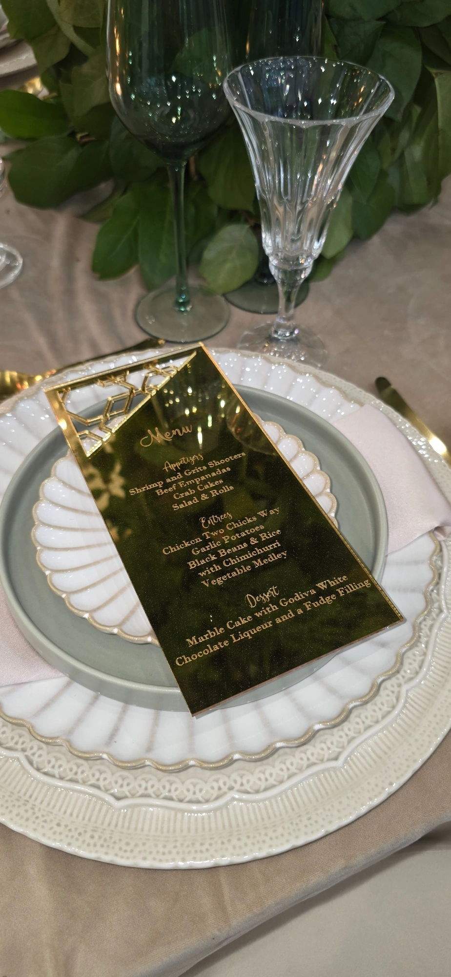 Gold menu, layered white plates, and crystal glassware beside greenery